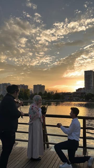 Gün batımında göl kenarındaki iskelede diz çökerek evlilik teklifi eden damat adayı ve keman çalan müzisyen
